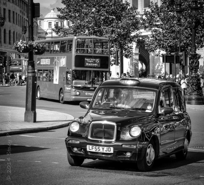 Black and white image of London taxi in foreground, London double-decker bus in background, entering a roundabout.